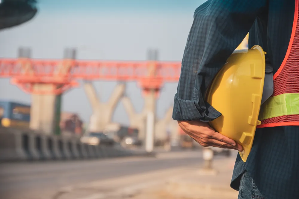Inspector con chaleco reflectante sosteniendo su casco amarillo frente a un viaducto en construcción